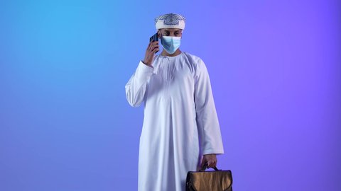 استخدام الأجهزة الحديثة والتقنيات المتطورة، مفهوم ادارة الاعمال عن بعد، مهن ووظائف مكتبيه عمانية، بورتريه لرجل عربي خليجي عماني يرتدي الدشداشة والكمه العمانية يمسك حقيبة و يقوم باجراء مكالمة هاتفية عبر الجوال، ارتداء الكمامة لإجراءات الوقاية من فايروس كورونا، خلفية زرقاء