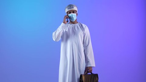 مفهوم ادارة الاعمال عن بعد، استخدام الأجهزة الحديثة والتقنيات المتطورة، مهن ووظائف مكتبيه عمانية، بورتريه لرجل عربي خليجي عماني يرتدي الدشداشة والكمه العمانية يمسك حقيبة و يقوم باجراء مكالمة هاتفية عبر الجوال، ارتداء الكمامة لإجراءات الوقاية من فايروس كورونا، خلفية زرقاء