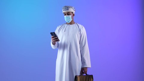 Browsing social media, communicating with family and friends, using modern technical devices, checking on relatives and hearing their news, portrait of an Arab Gulf Omani man wearing a dishdasha and a kummah, holding a mobile phone and bag in his hand, wearing a mask as preventive measures against the Corona virus, blue background