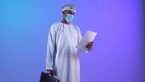 اعداد التقارير الخاصة بالعمل، بورتريه لشاب عربي خليجي عماني يرتدي الدشداشة و الكمة يحمل بيده مجموعة من الاوراق البيضاء و الحقيبة، مراجعة ومتابعة الخطط والمشاريع المكتبية بإيماءات التركيز، التدقيق في قراءة الملاحظات،ارتداء الكمامة لإجراءات الوقاية من فايروس كورونا، خلفية زرقاء