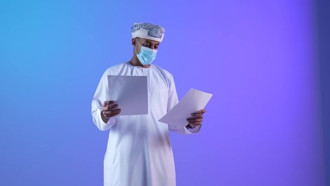 مفهوم التعلم الذاتي، القراءة والمطالعة ودراسة المشروع، بورتريه لرجل عربي خليجي عماني يرتدي الدشداشة والكمة يمسك بيده مجموعة أوراق يقوم بتصفحها بإيماءات التركيز والتمعن، قضاء وقت الفراغ في التثقيف والتعليم، ارتداء الكمامة لإجراءات الوقاية من فايروس كورونا، خلفية زرقاء
