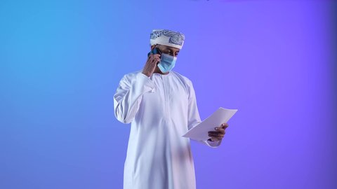 استخدام الأجهزة الحديثة والتقنيات المتطورة، مفهوم ادارة الاعمال عن بعد، مهن ووظائف مكتبيه عمانية، بورتريه لرجل عربي خليجي عماني يرتدي الدشداشة والكمه العمانية يمسك بيدة اوراق و يقوم باجراء مكالمة هاتفية عبر الجوال، ارتداء الكمامة لإجراءات الوقاية من فايروس كورونا، خلفية زرقاء