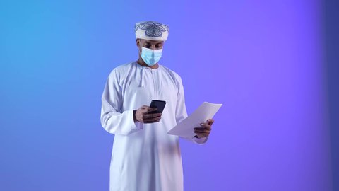 تسجيل الملاحظات والمعلومات المهمة،  بورتريه لشاب عربي خليجي عماني يرتدي الدشداشة والكمة يمسك بيده الهاتف المحمول، رتداء الكمامة لإجراءات الوقاية من فايروس كورونا، خلفية زرقاء
