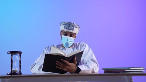 حب المطالعة والقراءة، تمضية أوقات الفراغ بالتعلم والتثقيف،الاهتمام بتصفح الكتب المفيدة، بورتريه مقرب لرجل عربي خليجي عماني يرتدي الدشداشه و الكمة يحمل بيده كتاب ، ارتداء الكمامة لإجراءات الوقاية من فايروس كورونا، خلفية زرقاء