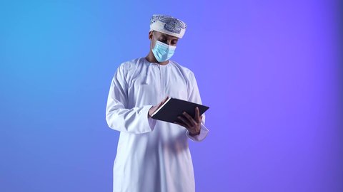 استخدام التكنولوجيا و التقنيات الحديثة, تصفح مواقع التواصل الاجتماعي، متابعة الأفلام والمسلسلات الممتعة، المحادثة مع الأهل والأصدقاء، بورتريه مقرب لرجل عربي خليجي عماني يرتدي الدشداشة و الكمة يحمل بيده جهاز التابلت،ارتداء الكمامة لإجراءات الوقاية من فايروس كورونا، خلفية زرقاء