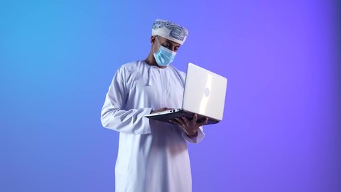 مهن ووظائف مكتبيه عمانية، بورتريه لرجل عربي خليجي عماني يرتدي الدشداشة والكمه العمانية  يعمل على جهاز الحاسوب المحمول بتركيز، ارتداء الكمامة لإجراءات الوقاية من فايروس كورونا، خلفية زرقاء