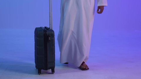 Expressions of walking and going to the airport, portrait of a smiling Omani Gulf Arab man wearing a dishdasha and a cloak holding a luggage bag, gestures of thinking and farewell, the concept of tourism and leisure trips, wearing a mask as a precaution against the Corona virus, blue background