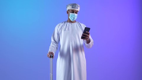 , الاستعداد لقضاء رحلة صيفية ممتعة، القيام بالجولات الترفيهية، بورتريه لشاب عربي عماني يرتدي الدشداشة و الكمة يقوم بتصفح الهاتف المحمول وبجانبه حقيبة الامتعة، ارتداء الكمامة لإجراءات الوقاية من فايروس كورونا, خلفية زرقاء
