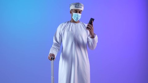 الاتفاق مع الاصدقاء على موعد الانطلاق, الاستعداد لقضاء رحلة صيفية ممتعة، القيام بالجولات الترفيهية، بورتريه لشاب عربي عماني يرتدي الدشداشة و الكمة يقوم بإجراء مكالمة هاتفية وبجانبه حقيبة الامتعة، ارتداء الكمامة لإجراءات الوقاية من فايروس كورونا, خلفية زرقاء