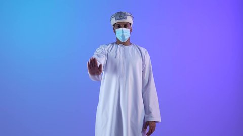 Expressions and signs to stop and not approach, portrait of a young Arab Gulf Omani man wearing a dishdasha and a kumrah, raising his hand up, looking at the camera with gestures of seriousness and firmness, refraining from doing something, wearing a mask as preventive measures against the Corona virus, blue background