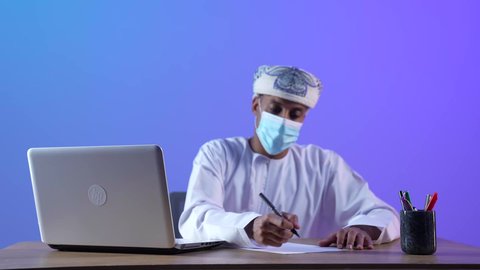 مفهوم ادارة الأعمال عن بعد، استخدام جهاز تقني حديث ومتطور، بورتريه لرجل عربي خليجي عماني يرتدي الدشداشة والكمة يقوم بكتابة شيء ما، دمج التكنولوجيا بالعمل، إنجاز المهام المطلوبة عبر اللابتوب، خلفية زرقاء