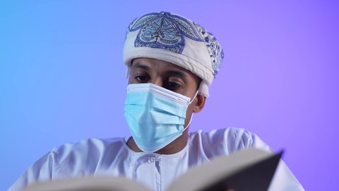 الاهتمام بتصفح الكتب المفيدة، تمضية أوقات الفراغ بالتعلم والتثقيف، حب المطالعة والقراءة، بورتريه مقرب لرجل عربي خليجي عماني يرتدي الدشداشه و الكمة يحمل بيده كتاب ، ارتداء الكمامة لإجراءات الوقاية من فايروس كورونا، خلفية زرقاء
