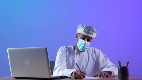 استخدام الأجهزة التقنية المتطورة، انجاز مهام الأعمال عن بعد، بورتريه مقرب لرجل عربي خليجي عماني يرتدي الدشداشة والكمه العمانية يجلس على المكتب و يعمل على جهاز الحاسوب المحمول و يكتب شيء ما، ارتداء الكمامة لإجراءات الوقاية من فايروس كورونا، خلفية زرقاء