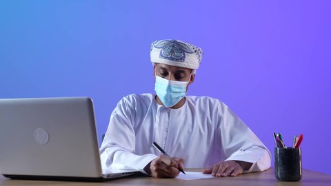 نجاز مهام الأعمال عن بعد، بورتريه مقرب لرجل عربي خليجي عماني يرتدي الدشداشة والكمه العمانية يجلس على المكتب و يعمل على جهاز الحاسوب المحمول و يكتب شيء ما، ارتداء الكمامة لإجراءات الوقاية من فايروس كورونا، خلفية زرقاء