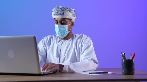 استخدام الأجهزة والتقنيات الحديثة، بورتريه مقرب لرجل عربي خليجي عماني يرتدي الدشداشة والكمه العمانية يجلس على المكتب و يعمل على جهاز الحاسوب المحمول بتركيز، ارتداء الكمامة لإجراءات الوقاية من فايروس كورونا، خلفية زرقاء