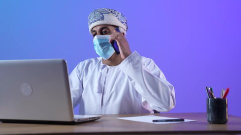 مهن ووظائف مكتبيه عمانية، مفهوم ادارة الاعمال عن بعد، استخدام الأجهزة الحديثة والتقنيات المتطورة، بورتريه مقرب لرجل عربي خليجي عماني يرتدي الدشداشة والكمه العمانية يجلس على المكتب و يقوم باجراء مكالمة هاتفية عبر الجوال، ارتداء الكمامة لإجراءات الوقاية من فايروس كورونا، خلفية زرقاء