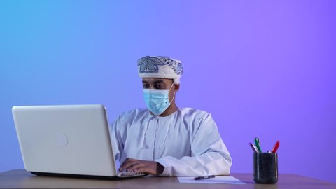 مهن ووظائف مكتبيه عمانية، بورتريه مقرب لرجل عربي خليجي عماني يرتدي الدشداشة والكمه العمانية يجلس على المكتب و يعمل على جهاز الحاسوب المحمول بتركيز، ارتداء الكمامة لإجراءات الوقاية من فايروس كورونا، خلفية زرقاء