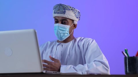 دمج التقنيات الحديثة والتكنولوجية بالمجال الإداري والمكتبية، بورتريه مقرب لرجل عربي خليجي عماني يرتدي الدشداشة والكمه العمانية يجلس على المكتب و يقوم باجراء مكالمه فيديو عبر الحاسب المحمول، ارتداء الكمامة لإجراءات الوقاية من فايروس كورونا، خلفية زرقاء