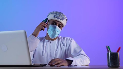 استخدام الأجهزة الحديثة والتقنيات المتطورة، مفهوم ادارة الاعمال عن بعد، مهن ووظائف مكتبيه عمانية، بورتريه لرجل عربي خليجي عماني يرتدي الدشداشة والكمه العمانية يجلس على المكتب و يقوم باجراء مكالمة هاتفية عبر الجوال، ارتداء الكمامة لإجراءات الوقاية من فايروس كورونا، خلفية زرقاء