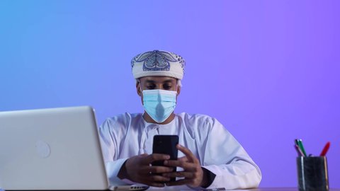 مفهوم ادارة الاعمال عن بعد، مهن ووظائف مكتبيه عمانية, استخدام الأجهزة الحديثة والتقنيات المتطورة، بورتريه لرجل عربي خليجي عماني يرتدي الدشداشة والكمه العمانية يجلس على المكتب و يقوم بتصفح الهاتف المحمول، ارتداء الكمامة لإجراءات الوقاية من فايروس كورونا، خلفية زرقاء