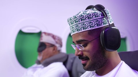 تصوير مقرب من الجانب لرجل عربي خليجي عماني يرتدي الدشداشه والكمه يجلس على كرسيه داخل الطائره يضع سماعة الرأس ويقوم بالإصغاء والاستماع إلى شيء ما، الاستماع الى الأغاني والموسيقى عبر سماعة الرأس، استخدام تقنية الهيدفون، طيران عمان