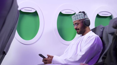 الاستماع الى الأغاني والموسيقى عبر سماعة الرأس، استخدام تقنية الهيدفون،تصوير مقرب من الجانب لرجل عربي خليجي عماني يرتدي الدشداشه والكمه يجلس على كرسيه داخل الطائره يضع سماعة الرأس ويقوم بالإصغاء والاستماع إلى شيء ما، طيران عمان
