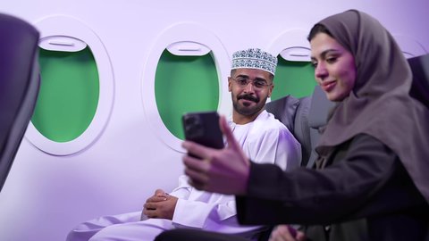 قضاء الاوقات السعيده داخل الطائره, مفهوم التسليه والترفيه وتبادل الاحاديث، تصوير مقرب من الجانب لرجل وامراه عربيان خليجيان عمانيين يرتديان الزي العماني يجلسان على كرسيهما داخل الطائره في رحله جويه يقومون بالتقاط صور سيلفي