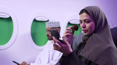 الاهتمام والعنايه بالمظهر الخارجي, التسويق لمستحضرات التجميل, تصوير مقرب من الجانب لامرأه عربيه خليجيه عمانيه ترتدي العباءة والحجاب تضع احداهما المكياج على وجهها داخل الطائره باستعمال فرشاة أحمر الخدود ، بجانبها رجل يستخدم الجوال