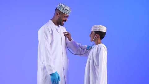 الفحص الطبي للأطفال ، الرعاية الطبية للأطفال ، تصوير لطبيب أطفال عربي خليجي عماني مع طفل صغير ، يضع الطفل سماعة الأذن يستمع إلى قلب الطبيب ، مفهوم الطب والرعاية الصحية