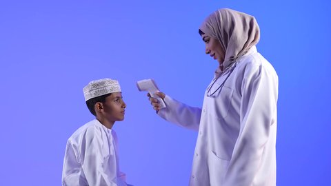 تصوير لطبيبة عربية عمانيه خليجية مع طفل ، طبيبة تحمل بيدها ميزان الحرارة الرقمي بالأشعة تحت الحمراء لقياس درجة الحرارة الجبهة من فايروس كورونا كوفيد-19 ، مفهوم الطب والرعاية الصحية ، مهن ووظائف طبية ، تقوم الطبيبة بفحص درجة حرارة الطفل