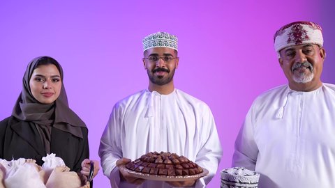 المودة والمحبة العائلية, تصوير لعائلة عربية خليجية عمانية تحتفل بالأعياد الإسلامية, رجل عماني يقوم بتقديم الحلويات التقليدية، طقوس الاحتفالات والمناسبات السعيدة، ضيافات الأعياد العربية الشعبية في سلطنة عمان، خلفية ملونة