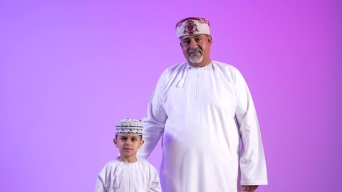 مشاعر الحب والمودة والأبوة، تصوير لرجل عربي خليجي عماني يرتدي الدشداشة والكمه يقوم بمعايدة ابنه في العيد، مفهوم بر الوالدين وتقديرهم، ارتداء الملابس العمانية التقليدية في الأعياد الإسلامية، خلفية ملونة