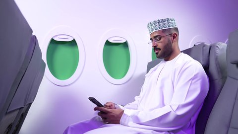 استخدام السياحة في إجازة الصيف, دمج التقنيات الحديثه فى الحياه اليوميه ، تصوير مقرب من الجانب لشاب عربي خليجي عماني يرتدي الدشداشه والكمه يجلس على كرسيه داخل الطائره ويمسك بيده هاتفهه المحمول