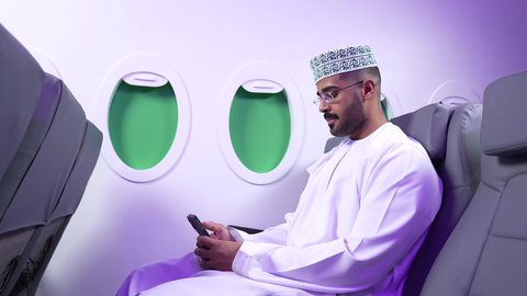 مفهوم ممارسه الانشطه والعمل عن بعد داخل الطائره, تصوير مقرب من الجانب لرجل عربي خليجي عماني يرتدي الدشداشه والكمه يجلس على كرسيه داخل الطائره الخليجيه يقوم بالعمل على جهاز الحاسوب المحمول, الطيران العماني الخليجي