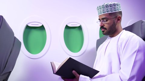 الطيران العماني الخليجي، ممارسه الانشطه الترفيهيه وهوايه المطالعه, تصوير مقرب من الجانب لرجل عربي خليجي عماني يرتدي الدشداشه والكمه يجلس على كرسيه داخل الطائره يقرأ كتابا بيده