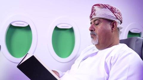 قراءه القصص والروايات, الاستمتاع وممارسه الهوايات المفضله ، الترفيه والتسليه وقضاء الاوقات داخل الطائره,  تصوير مقرب من الجانب لرجل عربي خليجي عماني يرتدي الزي العماني يجلس على كراسي الطائره في رحله جويه يقرأ الرجل كتابا