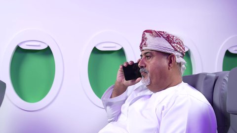 الاطمئنان على الاهل والاصدقاء ، اجراء مكالمه هاتفيه , تصوير مقرب من الجانب لرجل عربي خليجي عماني بدين يرتدي الدشداشه والكمه يجلس على كرسيه داخل الطائره الخليجيه يقوم باستخدام الهاتف المحمول, الطيران العماني الخليجي
