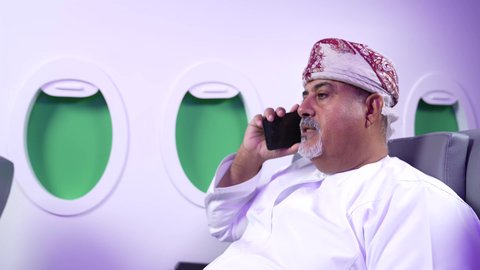 اجراء مكالمه هاتفيه , الاطمئنان على الاهل والاصدقاء ، تصوير مقرب من الجانب لرجل عربي خليجي عماني بدين يرتدي الدشداشه والكمه يجلس على كرسيه داخل الطائره الخليجيه يقوم باستخدام الهاتف المحمول, الطيران العماني الخليجي