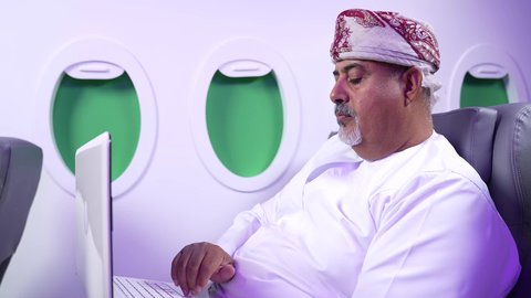 The concept of practicing activities and working remotely inside the plane, a close-up shot from the side of a fat Arab Gulf Omani man wearing a dishdasha and a kuma sitting on his chair inside the Gulf plane working on his laptop, Oman Gulf Airlines