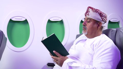 تصوير مقرب من الجانب لرجل عربي خليجي عماني بدين يرتدي الزي العماني يجلس على كراسي الطائره في رحله جويه يقرأ الرجل كتابا ،الترفيه والتسليه وقضاء الاوقات داخل الطائره, الاستمتاع بقرائه القصص والروايات, ممارسه الهوايات المفضله