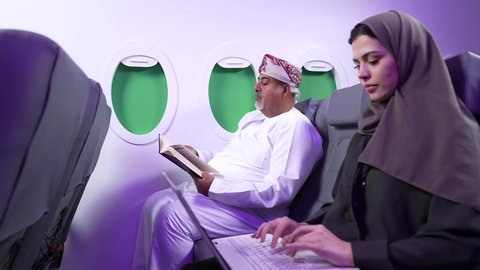 استخدام الحاسوب المحمول ، الاستمتاع بالقراءه والثقافه ، ممارسه الهوايات المفضله, الترفيه والتسليه وقضاء الاوقات داخل الطائره ، تصوير مقرب من الجانب لرجل بدين وامرأه عربيان خليجيان عمانيين يرتديان الزي العماني يجلسان على كراسي الطائره في رحله جويه