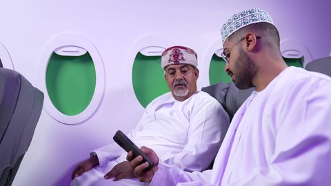 التسليه والترفيه والاندماج مع العائله، استخدام التكنولوجيا والتقنيات الحديثه ، قضاء الاوقات السعيده داخل الطائره, تصوير مقرب من الجانب لرجلان عربيان خليجيان عمانيين يرتديان الزي العماني يجلسان على كرسيهما داخل الطائره في رحله جويه