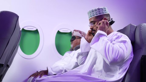 استخدام التكنولوجيا والتقنيات الحديثه ، الاستماع الى الموسيقي ، قضاء الاوقات السعيده داخل الطائره, التسليه والترفيه والاندماج مع العائله، تصوير مقرب من الجانب لرجلان عربيان خليجيان عمانيين يرتديان الزي العماني يجلسان على كرسيهما داخل الطائره في رحله جويه