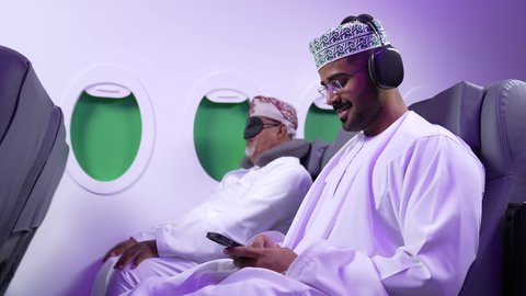 الاستماع الى الموسيقي ، استخدام التكنولوجيا والتقنيات الحديثه ، قضاء الاوقات السعيده داخل الطائره, التسليه والترفيه والاندماج مع العائله، تصوير مقرب من الجانب لرجلان عربيان خليجيان عمانيين يرتديان الزي العماني يجلسان على كرسيهما داخل الطائره في رحله جويه