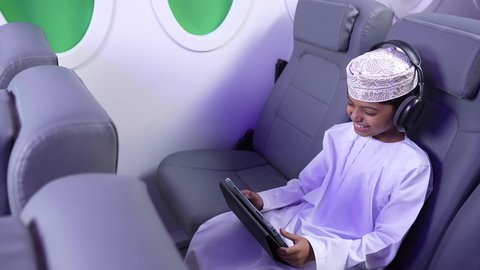اجراء مكالمه فيديو، إستخدام تقنيات الهيدفون، مفهوم الإصغاء والإستماع، قضاء اوقات ممتعة ومسلية، تصوير من الامام لطفل مسافر عربي خليجي عماني يرتدي الدشداشة و الكمه مبتسم يجلس في مقعد الطائرة يمسك بيد التابلت، رحلة سياحية من خلال خطوط الطيران العماني