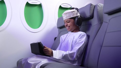 إستخدام تقنيات الهيدفون، مفهوم الإصغاء والإستماع، قضاء اوقات ممتعة ومسلية، تصوير من الجانب لطفل مسافر عربي خليجي عماني يرتدي الدشداشة و الكمه مبتسم يجلس في مقعد الطائرةيمسك بيد التابلت، رحلة سياحية من خلال خطوط الطيران العماني