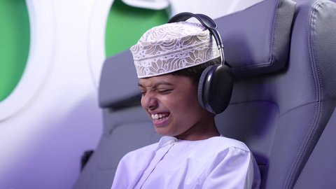 رحلة سياحية من خلال خطوط الطيران العماني، مفهوم الإصغاء والإستماع، إستخدام تقنيات الهيدفون، استخدام التكنولوجيا و التقنيات الحديثة المتطورة، تصوير مقرب لطفل مسافر عربي خليجي عماني يرتدي الدشداشة و الكمه مبتسم يجلس في مقعد الطائر بايماءات السعادة و السرور