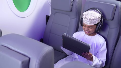 مفهوم الإصغاء والإستماع، قضاء اوقات ممتعة ومسلية، إستخدام تقنيات الهيدفون، استخدام التكنولوجيا و التقنيات الحديثة المتطورة، تصوير من الامام لطفل مسافر عربي خليجي عماني يرتدي الدشداشة و الكمه مبتسم يجلس في مقعد الطائرةيمسك بيد التابلت، رحلة سياحية من خلال خطوط الطيران العماني
