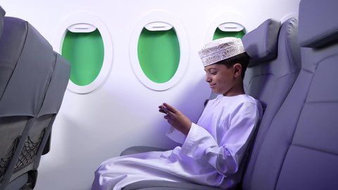 الاستعداد لقضاء عطلة صيفية ممتعة، مفهوم السياحة و السفر، استخدام التكنولوجيا و التقنيات الحديثة المتطورة، تصوير من الجانب لطفل مسافر عربي خليجي عماني يرتدي الدشداشة و الكمه مبتسم يجلس في مقعد الطائرة ويقوم باللعب على الهاتف المحمول، رحلة سياحية من خلال خطوط الطيران العماني