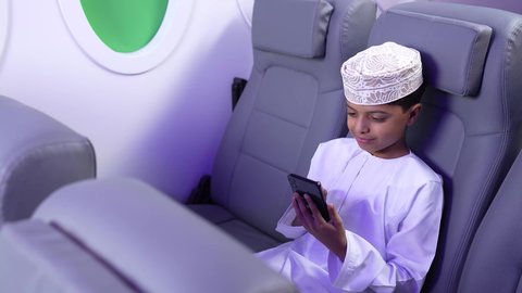 مفهوم السياحة و السفر، استخدام التكنولوجيا و التقنيات الحديثة المتطورة، الاستعداد لقضاء عطلة صيفية ممتعة، تصوير من الامام لطفل مسافر عربي خليجي عماني يرتدي الدشداشة و الكمه مبتسم يجلس في مقعد الطائرة و بتصفح الهاتف المحمول، رحلة سياحية من خلال خطوط الطيران العماني